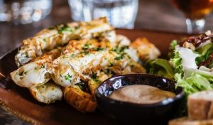 Closeup image of kingfish goujons served on a dark plate with a little dish of sauce and salad on the side.